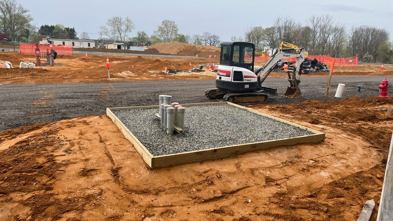Construction site with a concrete utility pad, excavator, and muddy ground under overcast sky