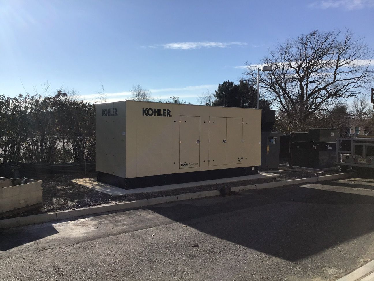 Large tan KOHLER generator unit beside a road under a blue sky, with trees and buildings nearby