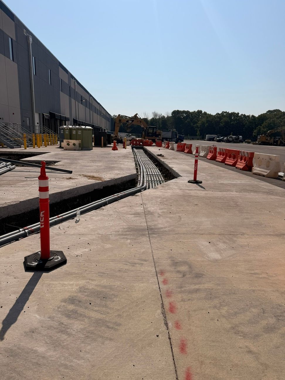 Construction area beside a building with orange cones, red barriers, and a newly poured concrete walkway under blue sky