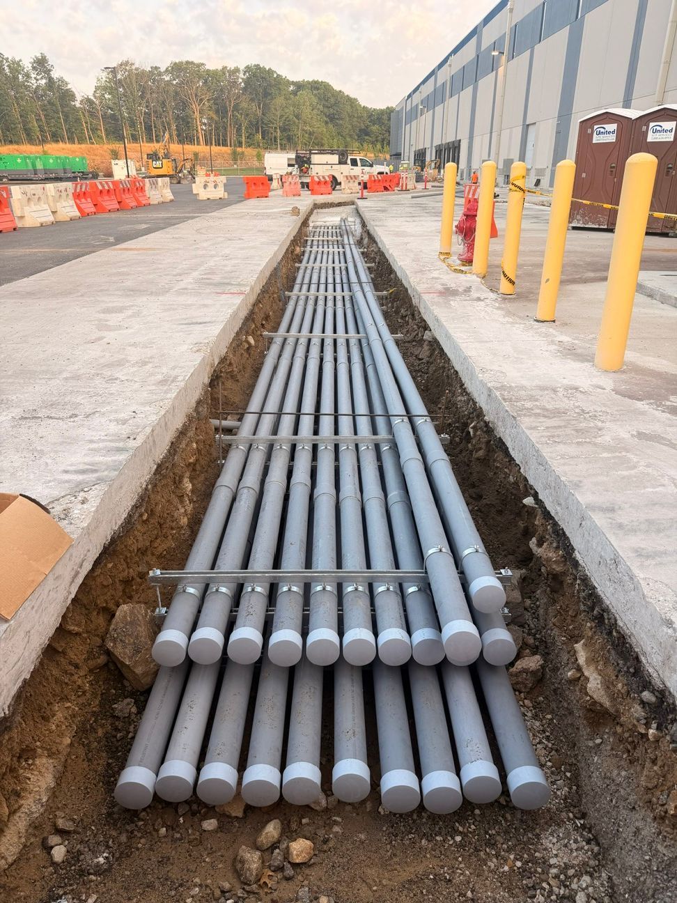 Gray utility pipes laid in a trench at a construction site beside a concrete road and yellow bollards