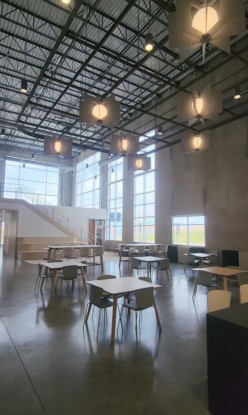 Bright cafeteria-style room with tables, chairs, large windows, and hanging lights under an exposed ceiling