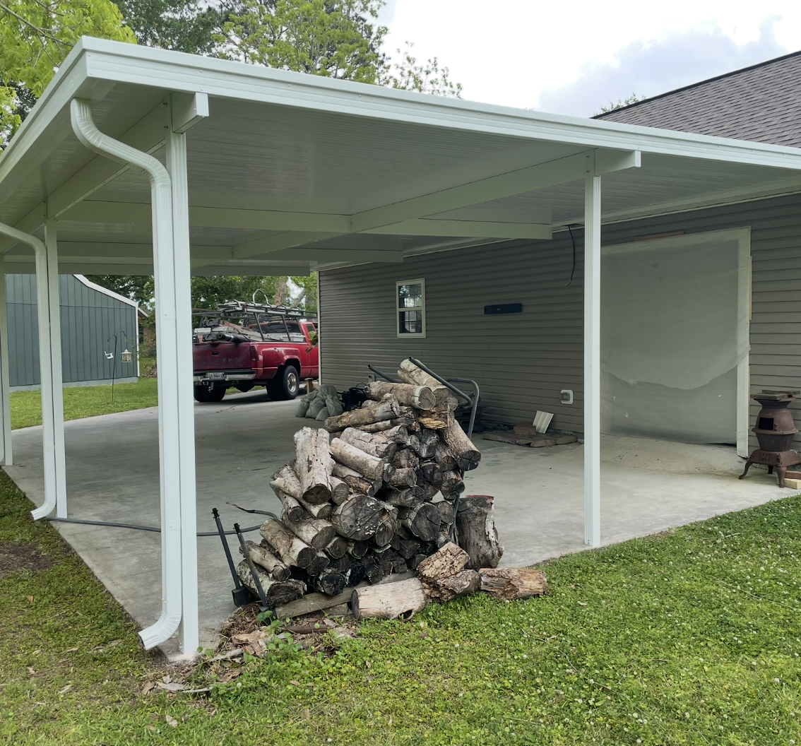 A red truck is parked under a covered carport next to a pile of logs.