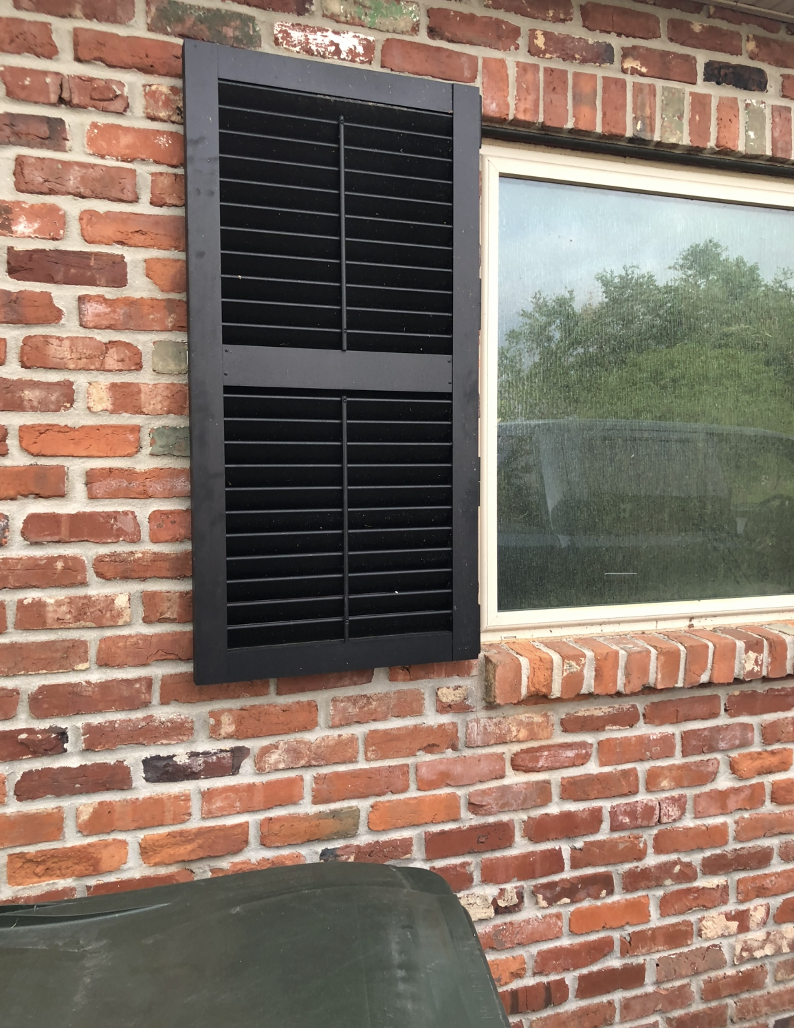 A black shutter is hanging on a brick wall next to a window.