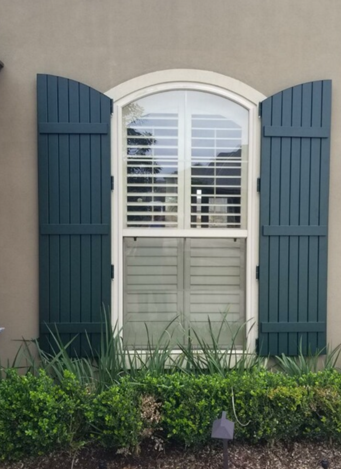 A window with blue shutters on a house with a hedge in front of it.