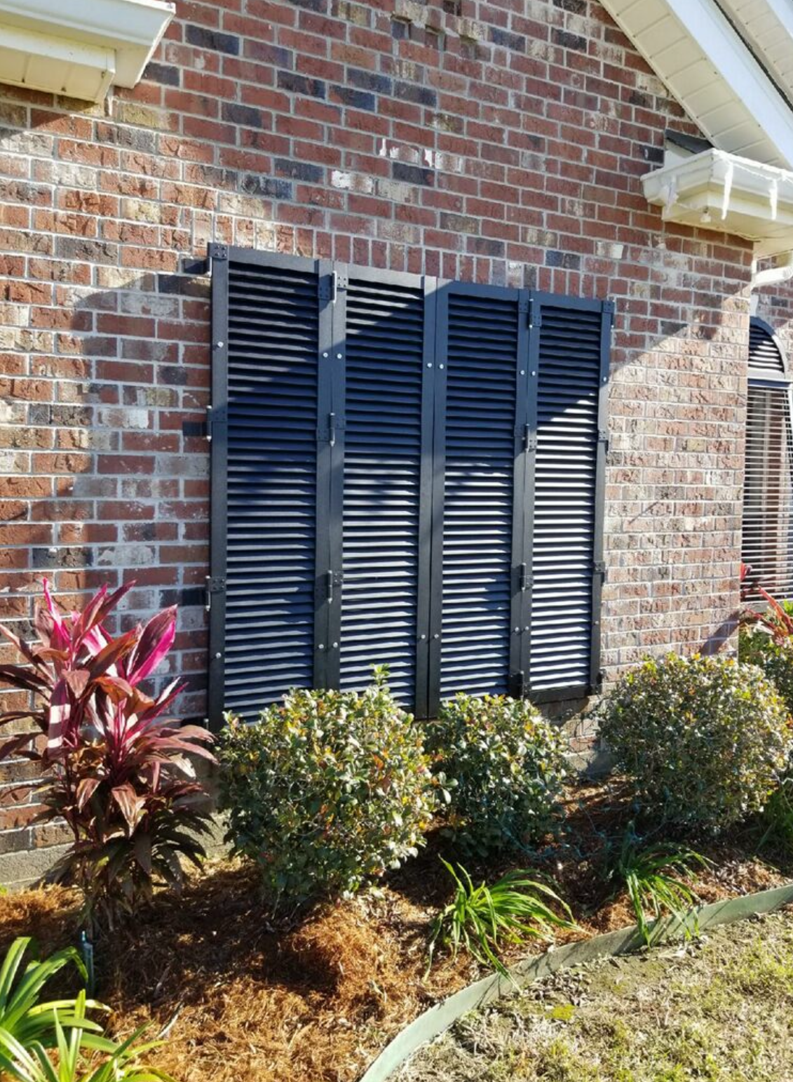 A brick building with shutters on the windows and bushes in front of it.