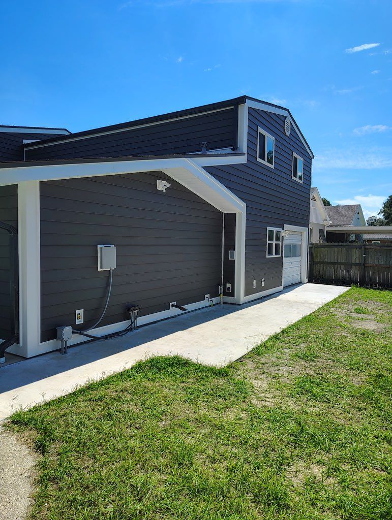 A two-story building with dark gray horizontal siding, white trim, and a concrete walkway beside a grass lawn.