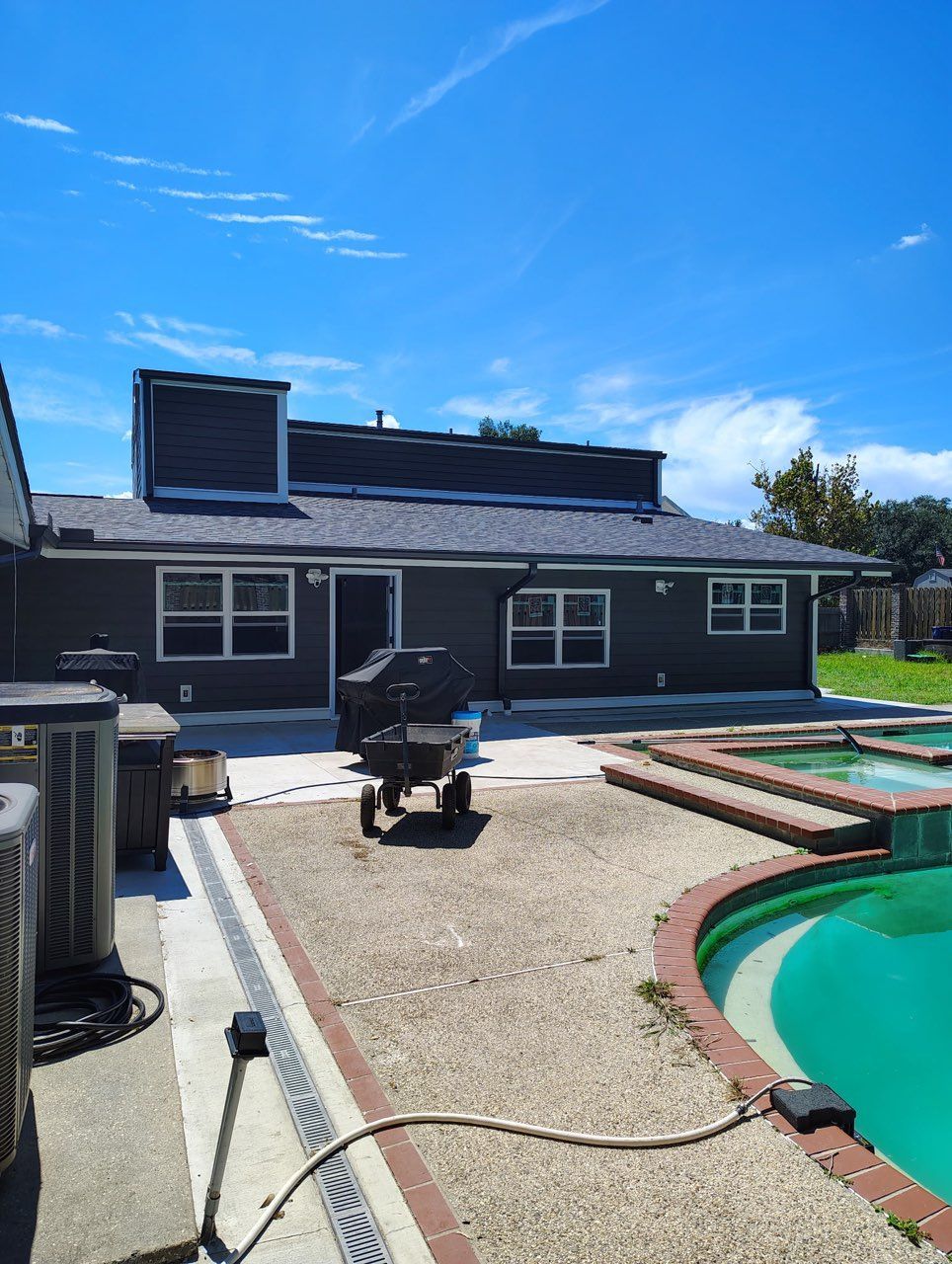 A dark gray single-story house with a patio, a pool, and a small equipment cart under a bright blue sky.