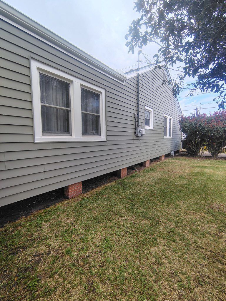 A side view of a gray, single-story house elevated on brick piers, with white-framed windows set against a grassy lawn.