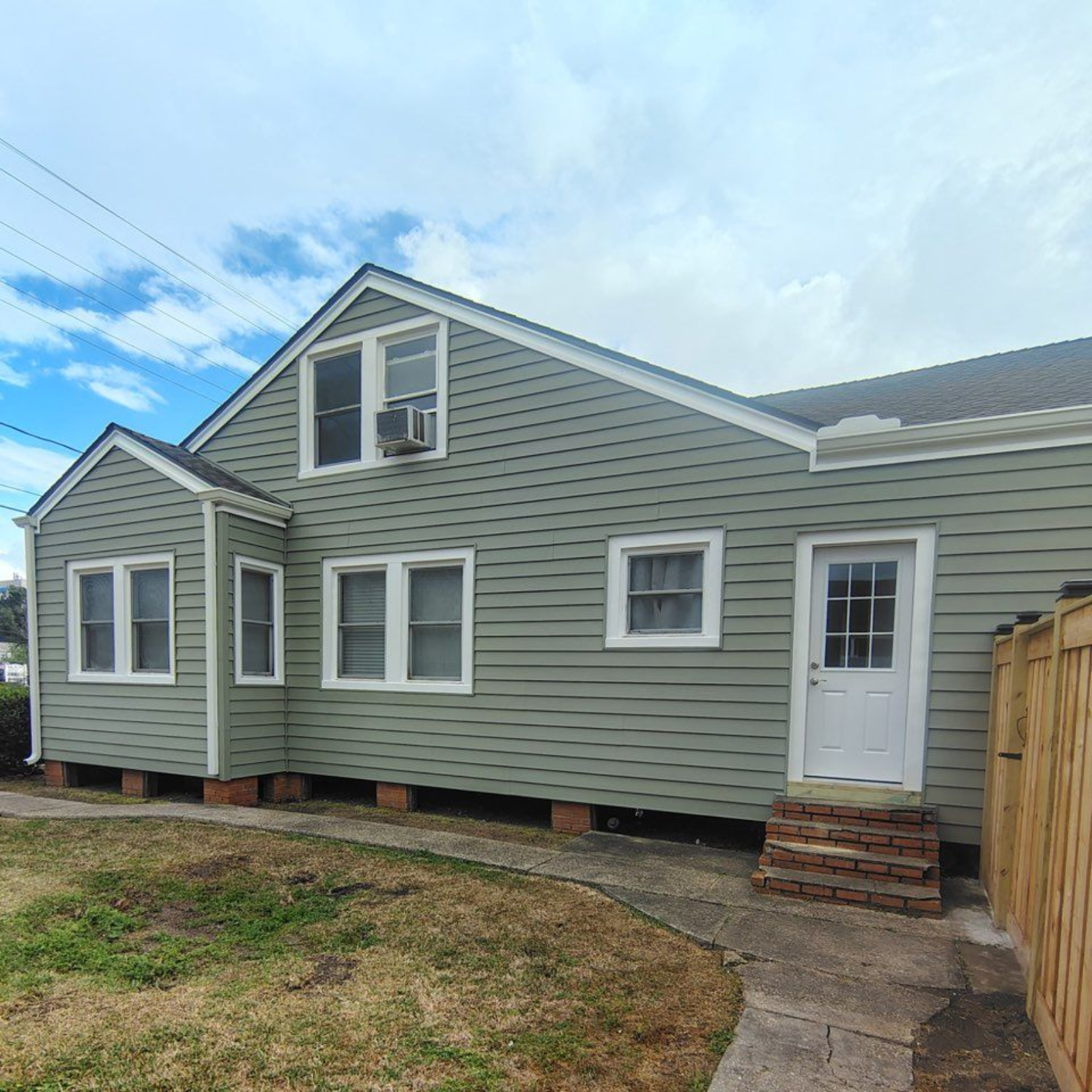 Light green, two-story house with white-trimmed windows, a brick foundation, and a side entry door, viewed from a lawn.
