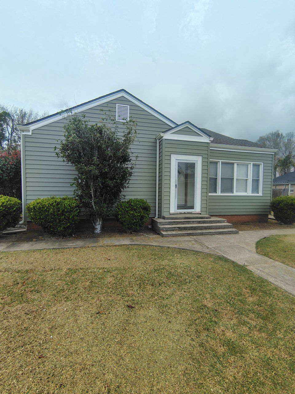 A one-story sage green house with a small front porch, a central door, large windows, and a manicured lawn.