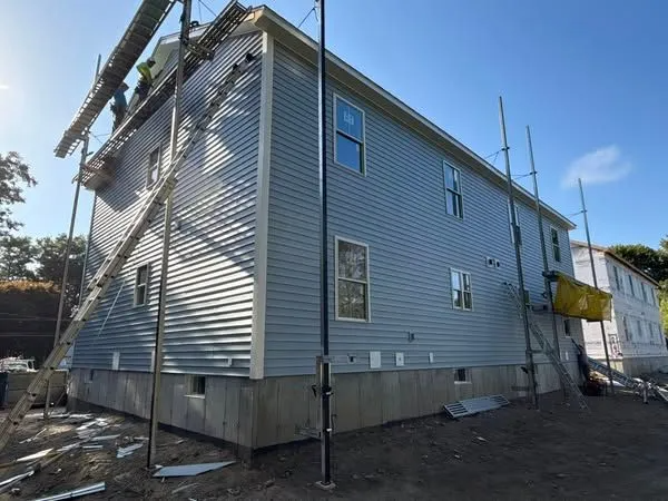 Two-story building with gray siding under construction, scaffolding, workers on the roof against a blue sky.