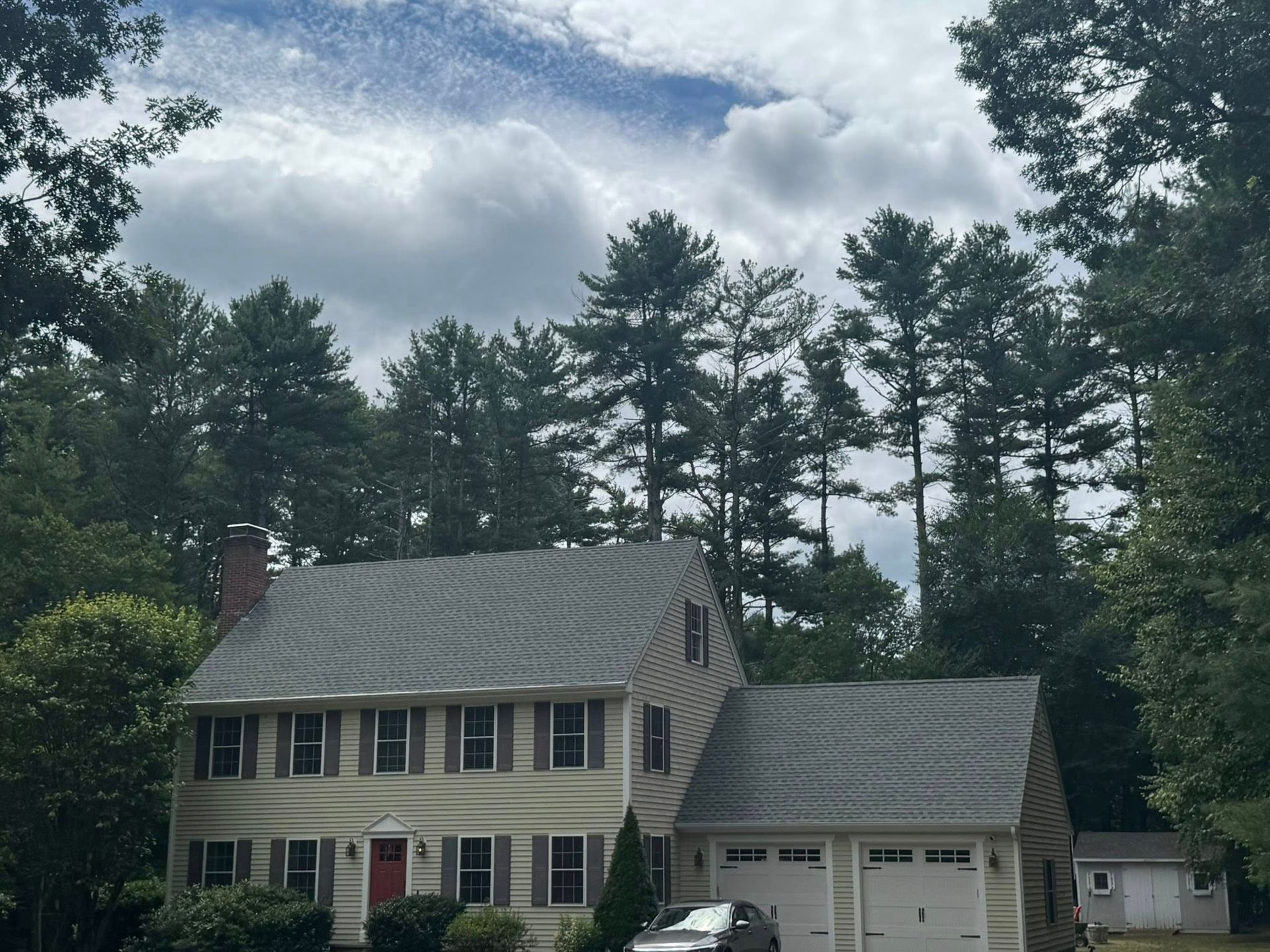Two-story beige house with gray roof and attached garage, tall trees in background, cloudy sky.