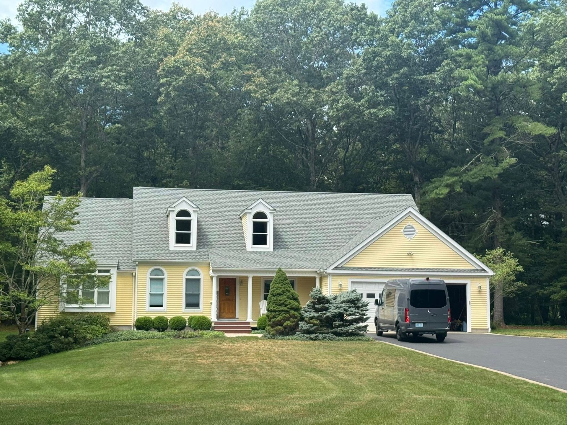 Yellow house with gray roof, two dormers, brown door, green lawn, trees, and a van in driveway.