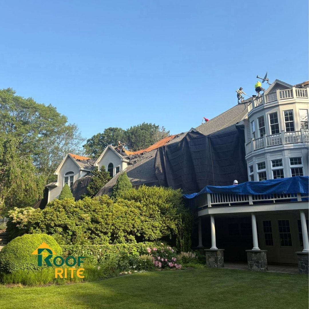 Roof repair on a large house with a tarp and workers. Green yard with trees and blue sky.