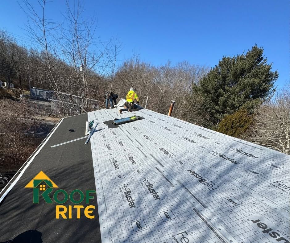 Workers installing silver roofing underlayment on a sloped roof with blue sky and trees in the background