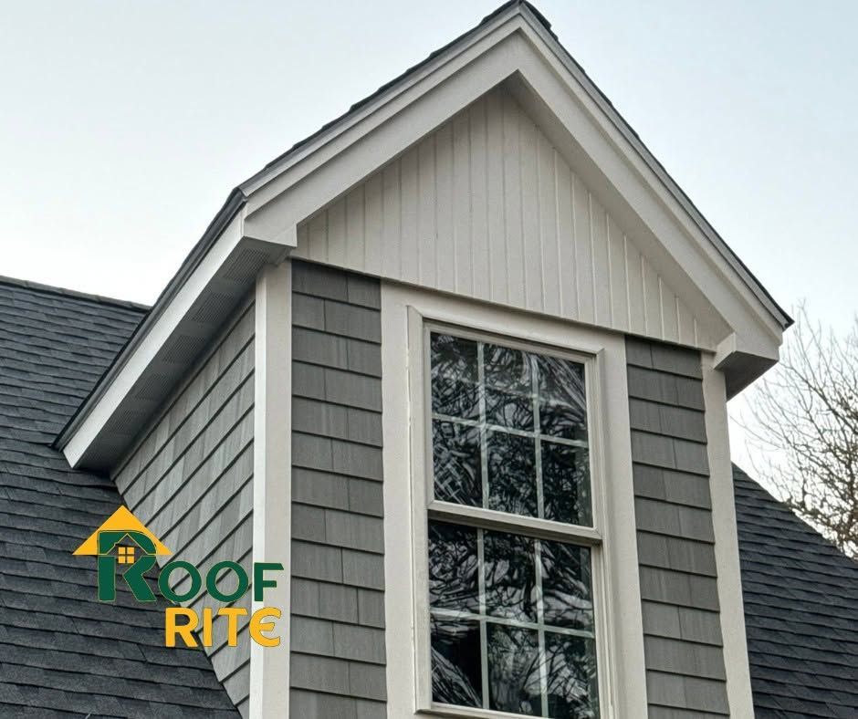 Gray house dormer with white trim and large window, roof repair company logo in lower left
