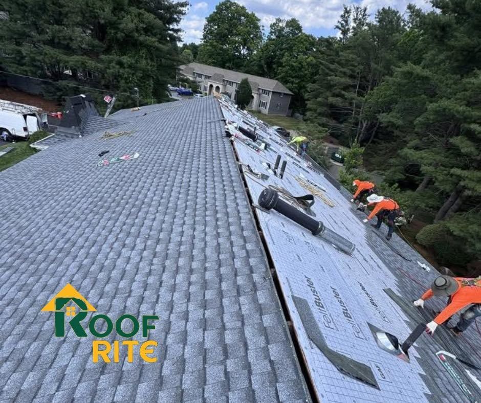 Roof workers installing underlayment on a gray shingle roof, with tools and a house in the background.