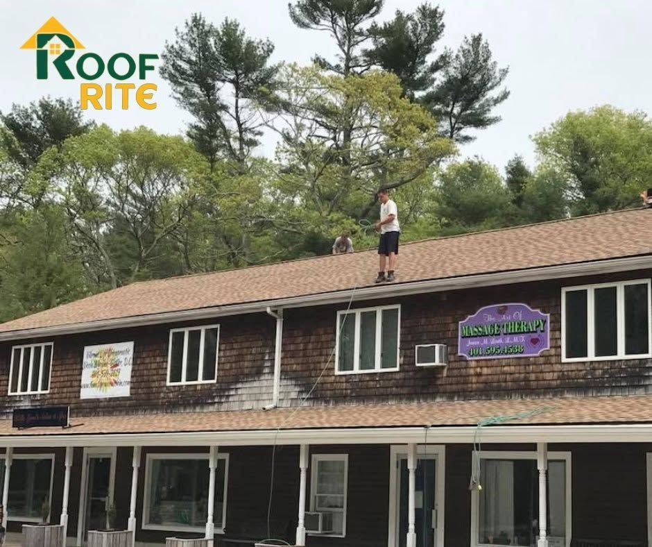 Worker on a shingle roof above a two-story building and trees behind.
