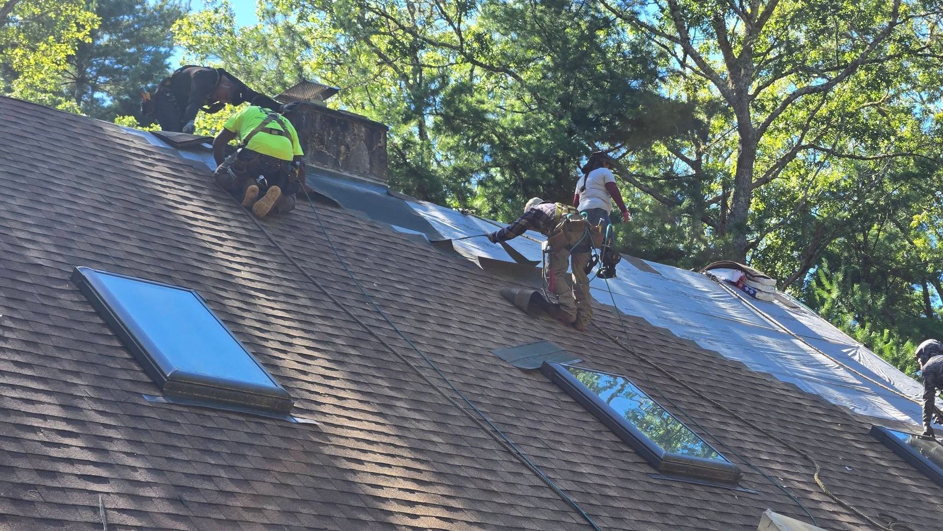 Workers installing solar panels on a steep shingled roof under trees