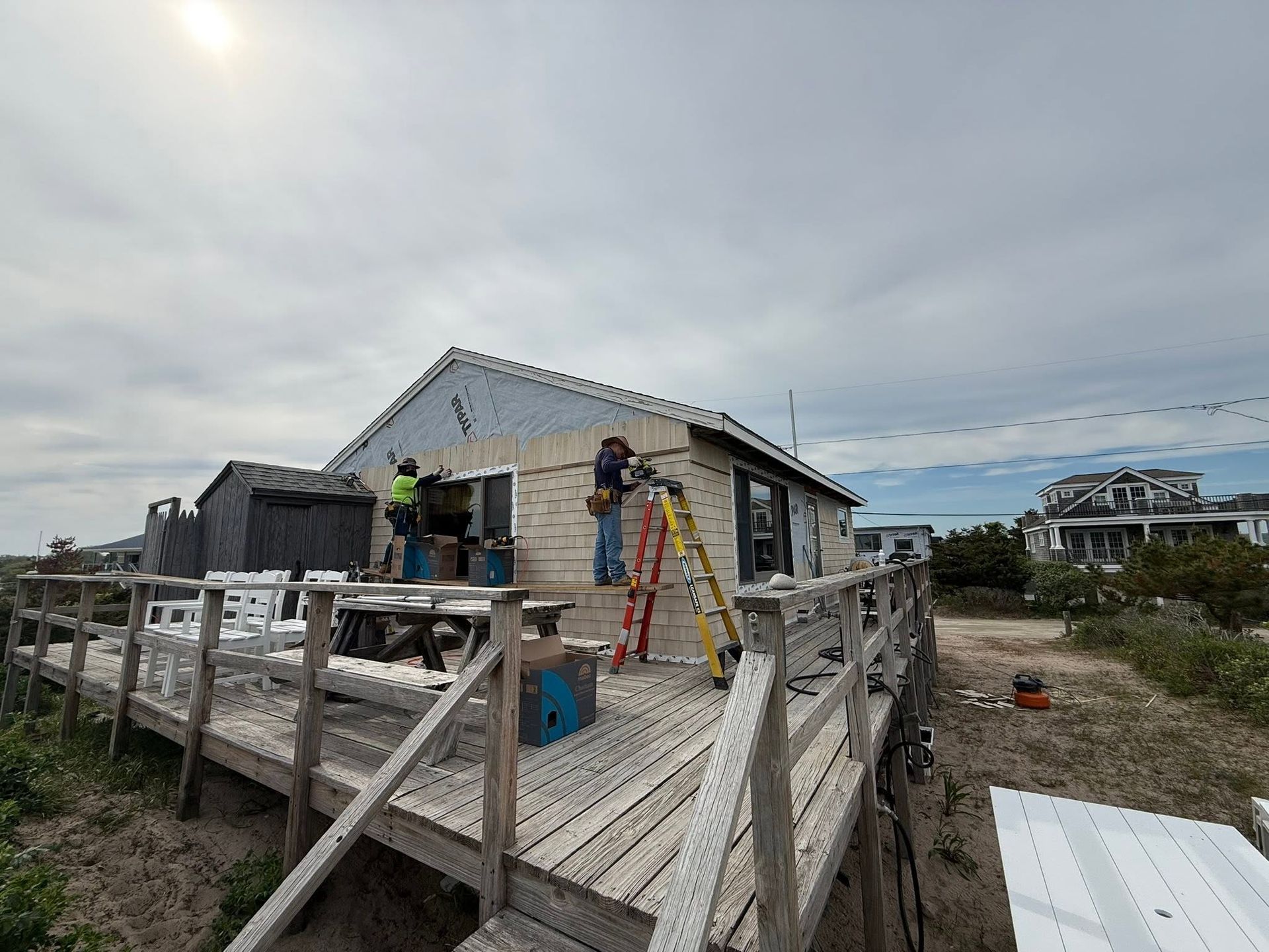 Workers on a raised wooden deck beside a light-colored house under construction, with ladders and tools visible.