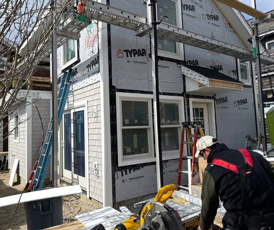 Workers on a wooden deck framing a house under construction, with ladders and scaffolding beside a cloudy sky.