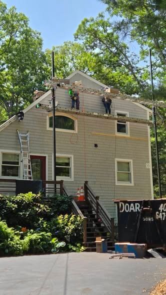 Two-story beige house with workers on the roof and a ladder leaning against the side, surrounded by trees.