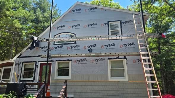 Two-story house under renovation with Tyvek wrap, windows, and ladders along the front and side.