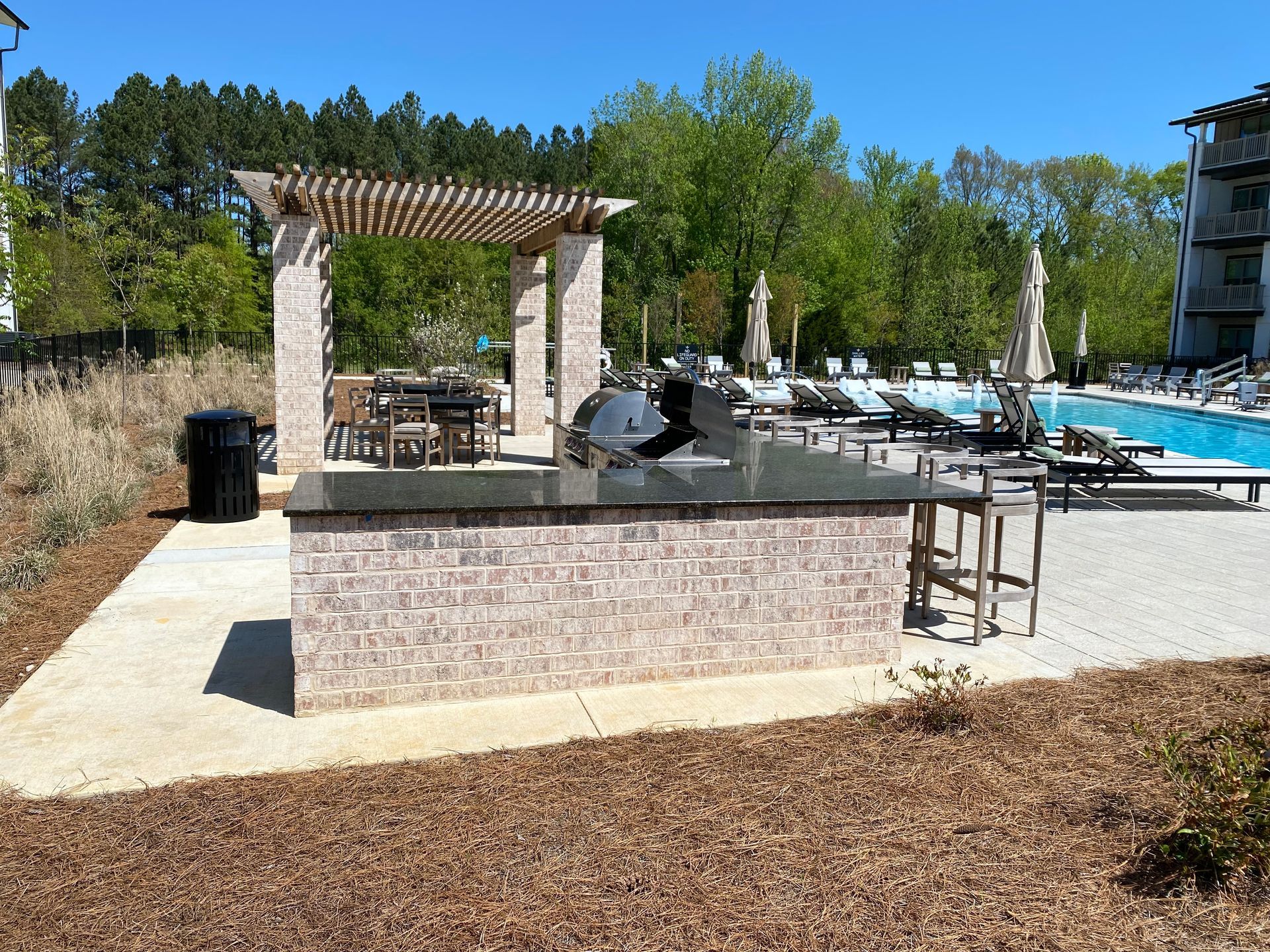 A grill is sitting under a pergola next to a pool.