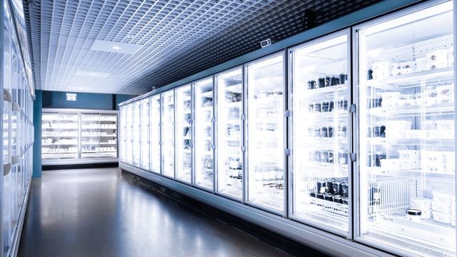 A long, brightly lit aisle in a grocery store featuring glass-fronted refrigerated display cases filled with products.