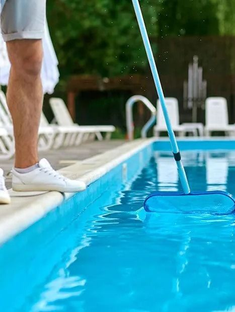 Person cleaning a blue swimming pool with a net, wearing white shorts and shoes.