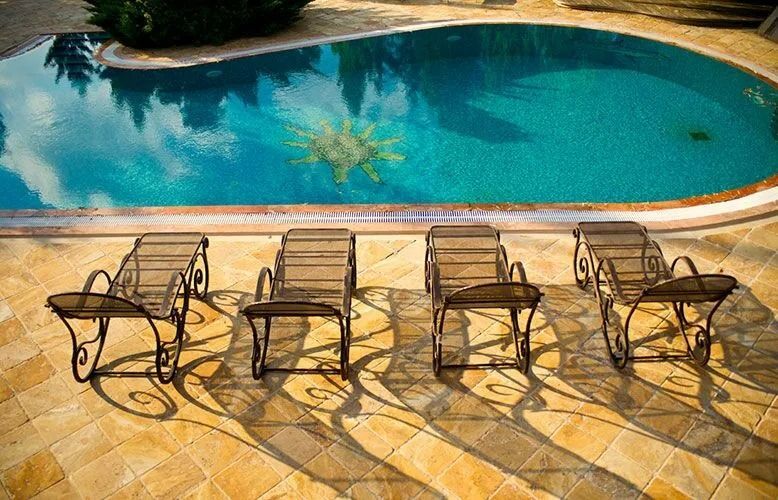 Lounge chairs by a turquoise pool reflect sky and trees; the pool deck is tan-colored stone.
