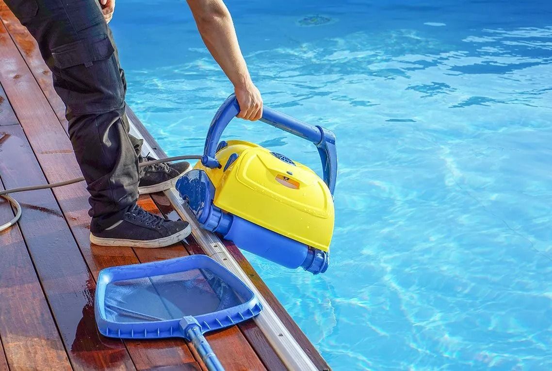 Person holding a yellow and blue robotic pool cleaner near a pool, with a blue skimmer nearby.