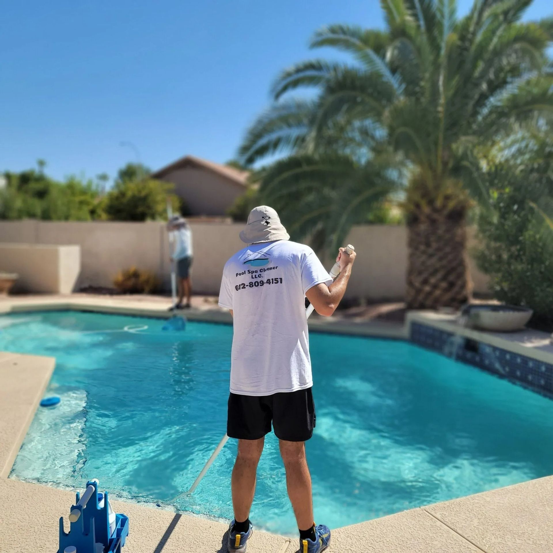 Two people cleaning a blue swimming pool on a sunny day. One wears a white shirt with a logo, the other a hat.