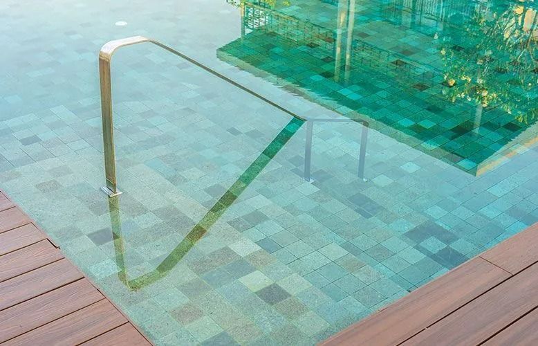 Pool with handrail and wooden deck, water reflecting green tile and foliage.