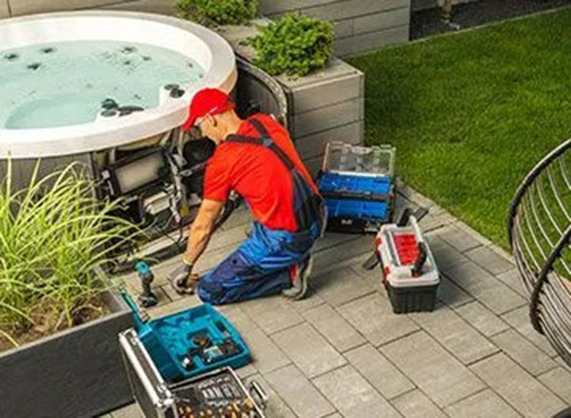 Man in overalls fixing a hot tub, surrounded by tools, on a patio next to a lawn.