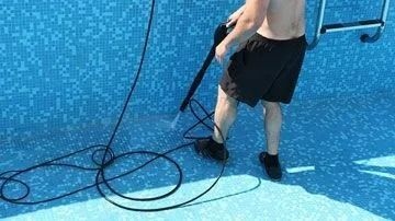 Person cleans a drained blue-tiled pool with a hose, standing near a ladder.