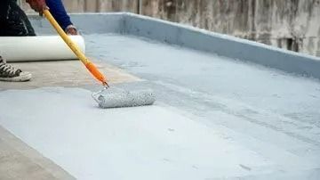 Person applying gray waterproof coating to a flat roof with a paint roller.
