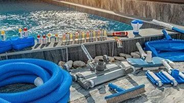 Pool cleaning tools on a pool deck: hoses, brushes, vacuums, test kits, and floats.