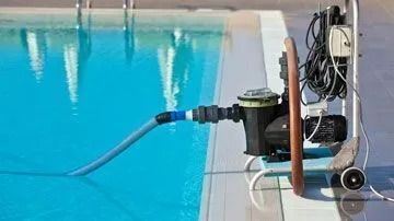 Pool pump system near a swimming pool, blue water, hose connected to the pump, and a white wheeled stand.