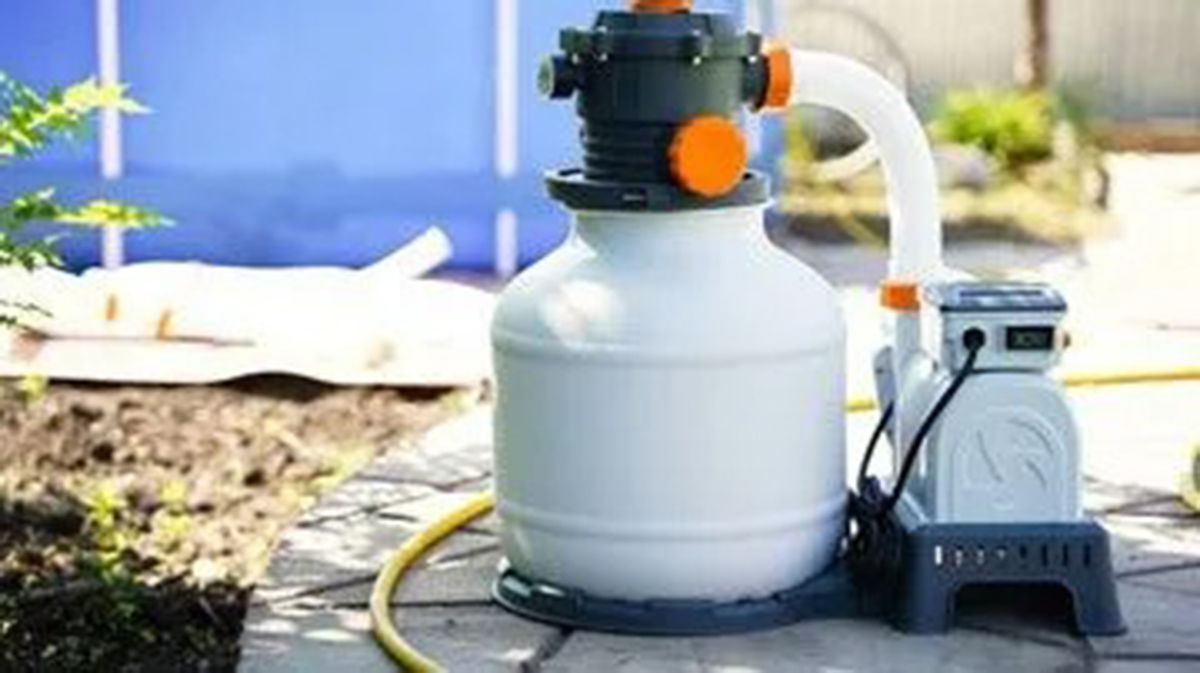 White and grey pool filter system outside near a blue pool, a hose, and greenery.