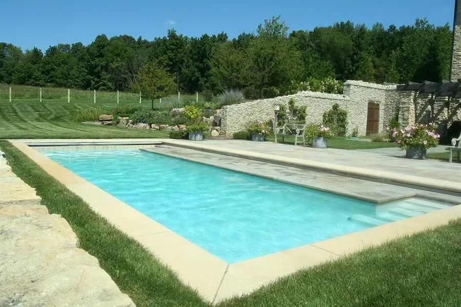 Rectangular outdoor pool with light blue water, surrounded by light stone and green grass. Forest in the background.