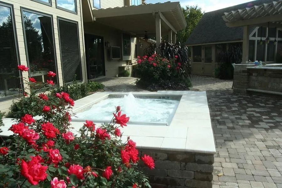 A patio with a water feature surrounded by red roses, brick and stone pavers, and a house in the background.