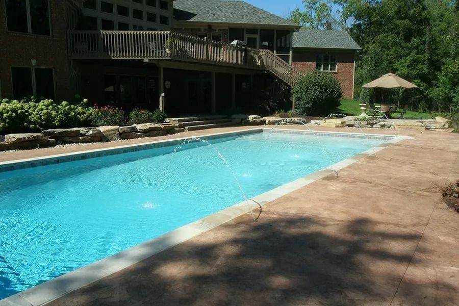 Long rectangular swimming pool in a backyard, beside a house. Brown patio, blue pool water, a small fountain is active.
