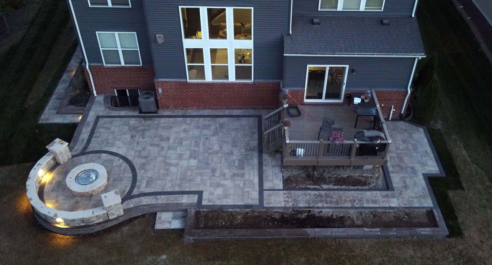 Aerial view of a home's backyard with a patio, fire pit, and deck lit up at dusk.