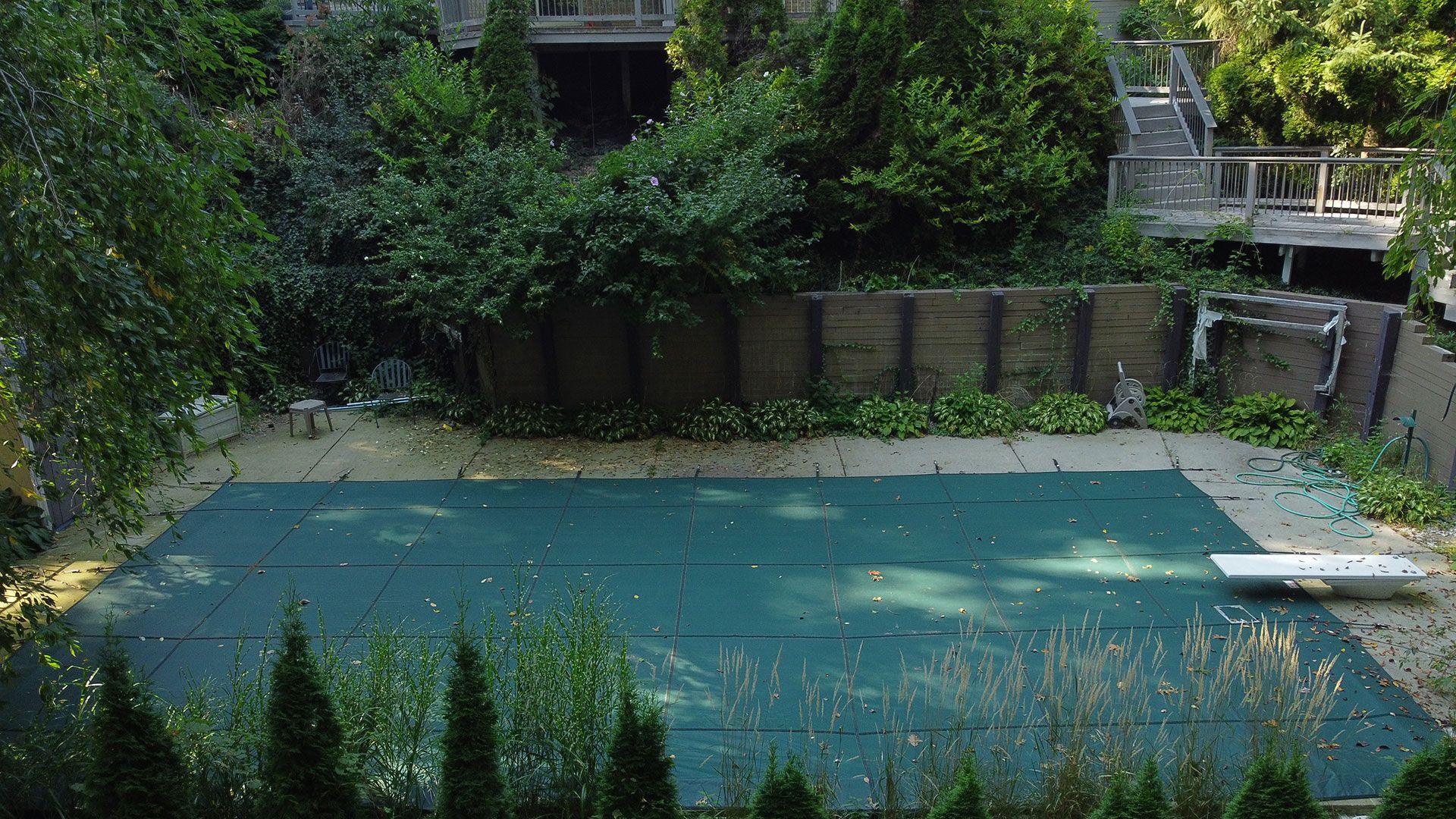 Pool covered with green tarp, surrounded by greenery and a deck in the background.
