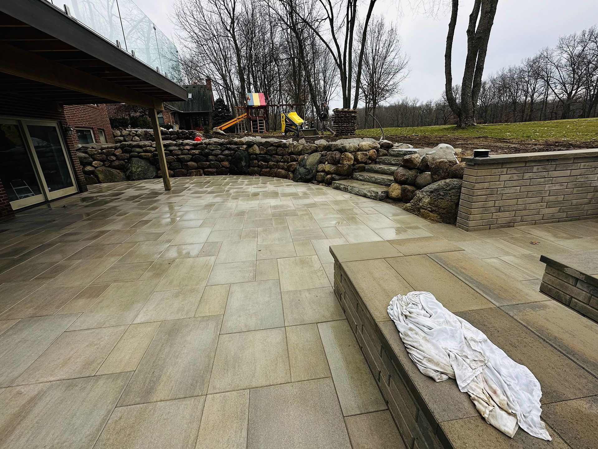 Stone patio with rock wall, glass railing, and playground in the background. A white cloth rests on a table.