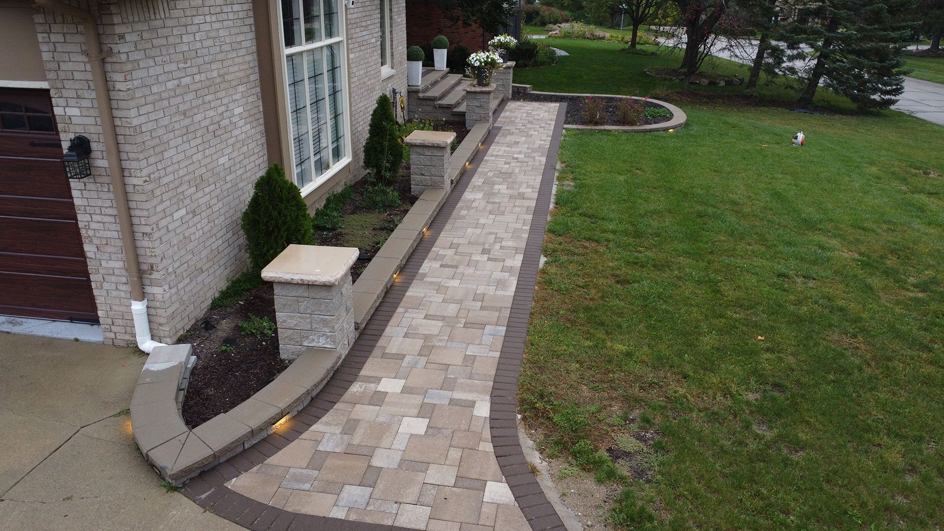 Brick pathway alongside a house with green lawn. Stone pillars and flowerbeds.
