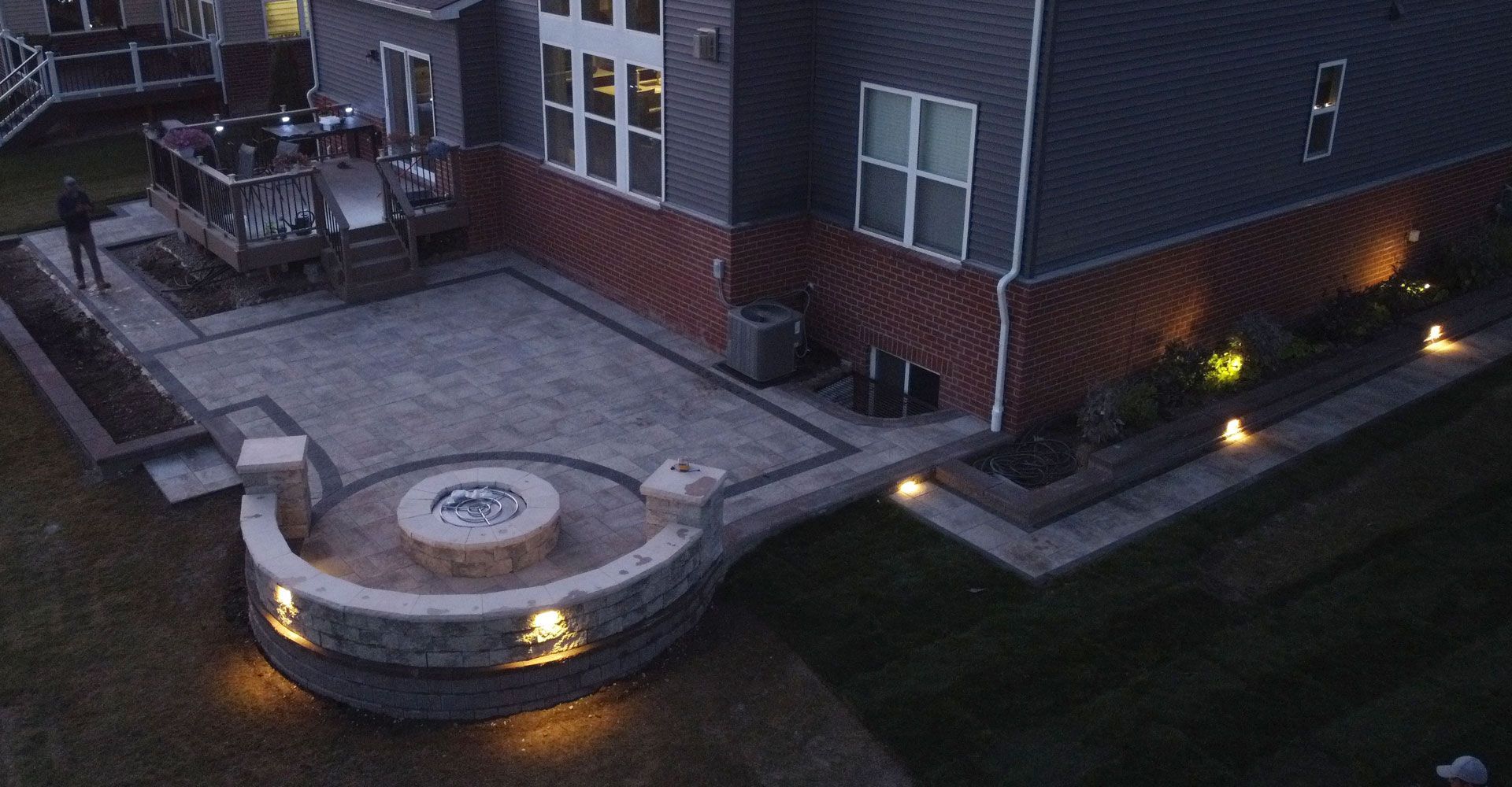 Night view of a backyard patio with a fire pit, lighting, and a deck attached to a house.