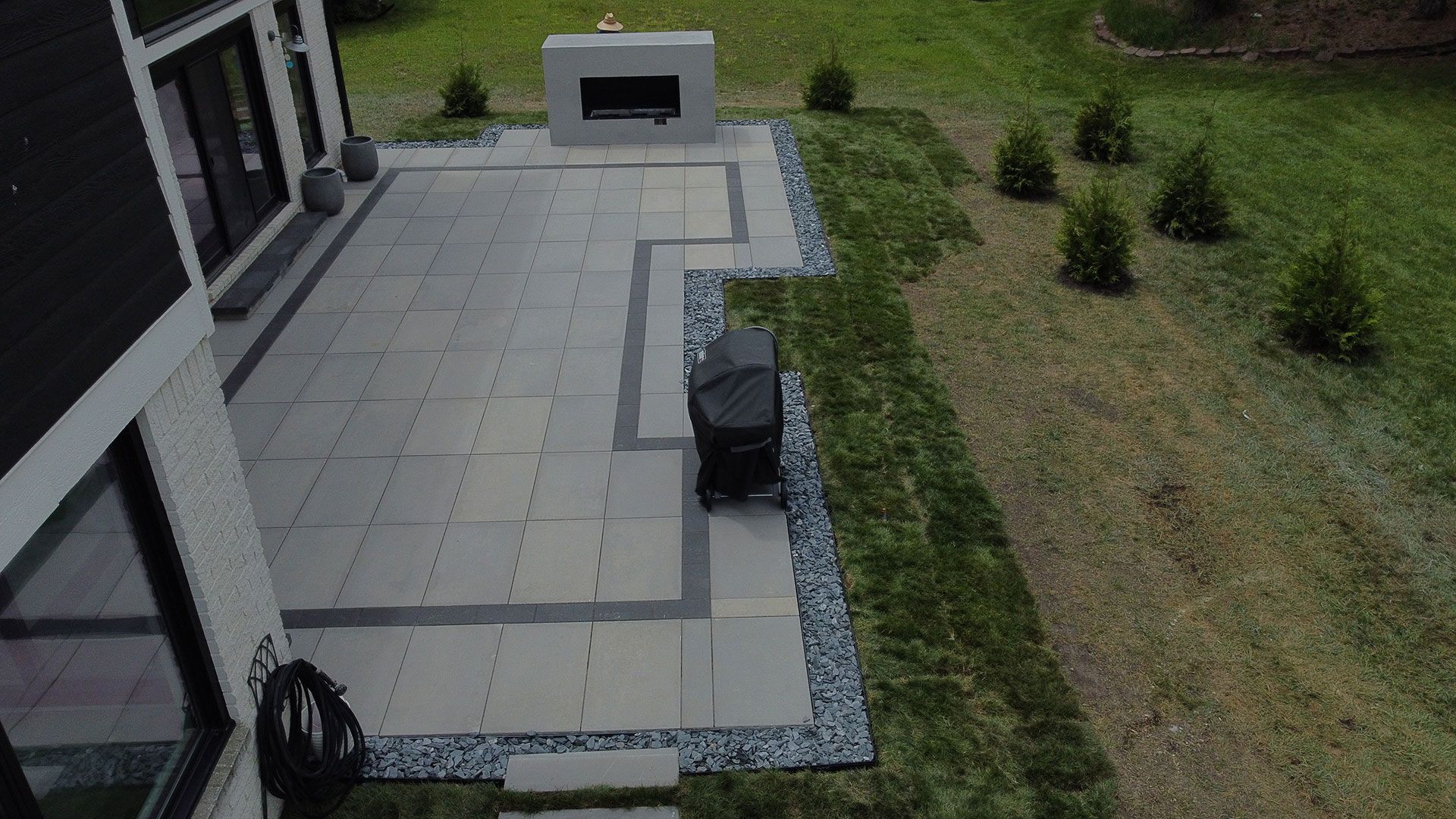 Outdoor patio with gray pavers, black trim, built-in fireplace, and grill; next to a lawn.