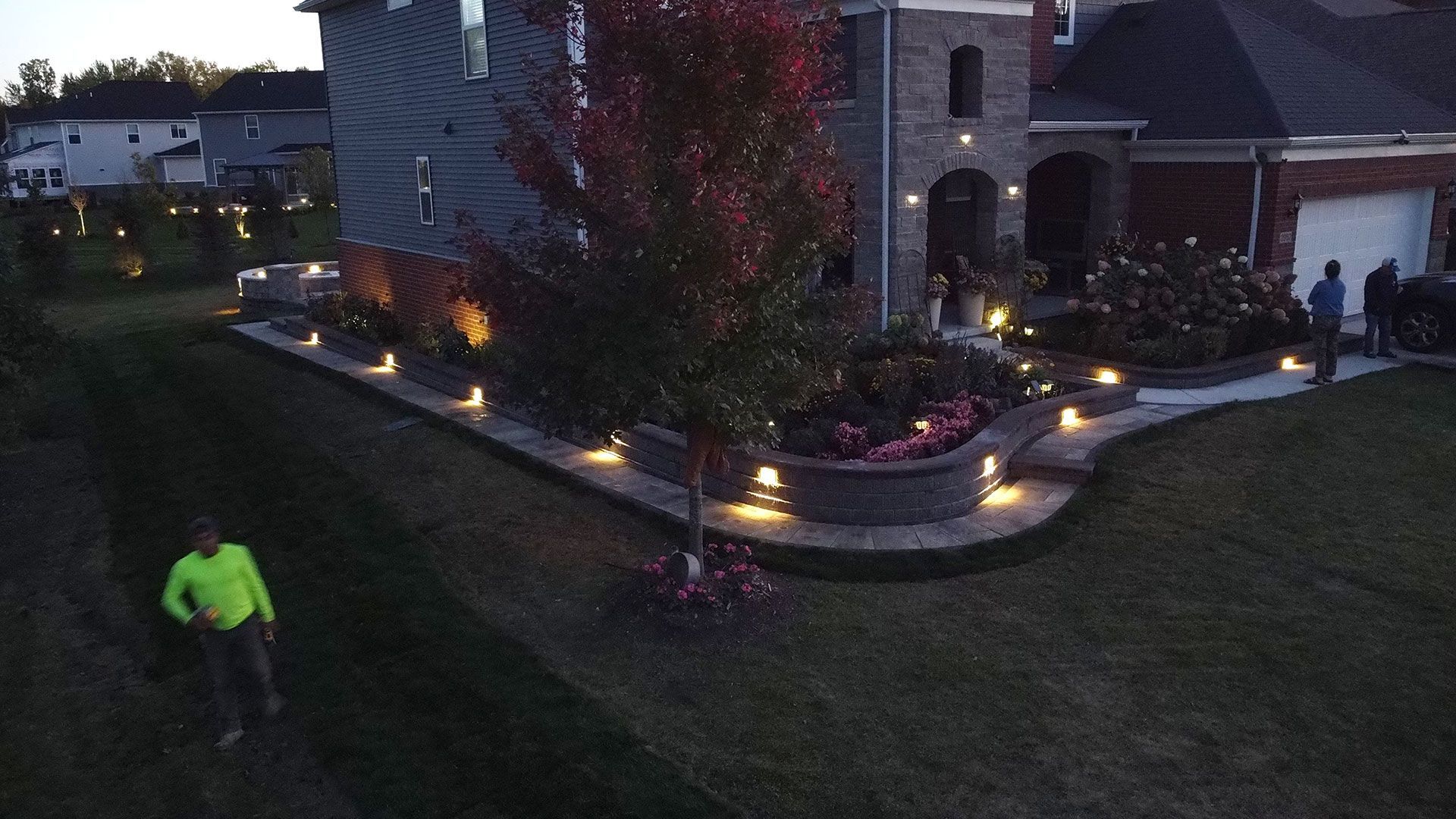 Evening view of a house with lit landscaping. A person walks on the grass.